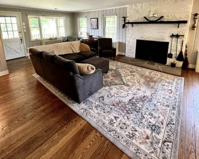 A living room with a light-colored sofa, two armchairs, a large area rug, and a stone fireplace against a white wall.