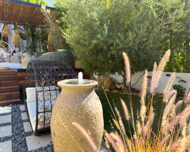 The image depicts a serene garden setting featuring a decorative stone fountain, a circular wicker chair, and lush greenery, including ornamental grasses and an olive tree.