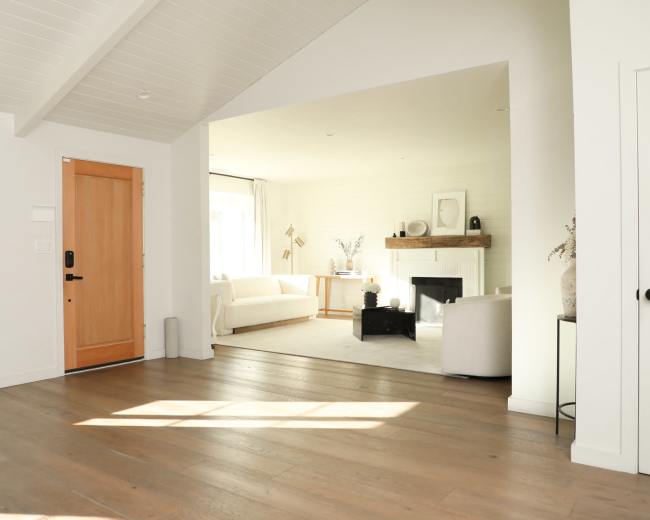 The image shows a bright, open interior space featuring a wooden front door, hardwood floors, and a living area with a white sofa and a fireplace.