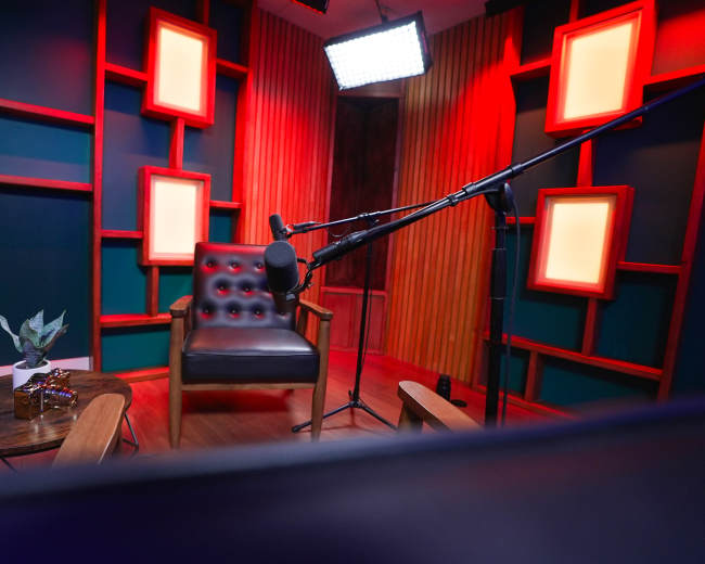 A neatly arranged podcast or interview setup featuring a black chair, a microphone on a stand, and a coffee table with a small plant and decorative items, all set against a backdrop of illuminated frames and colorful walls.