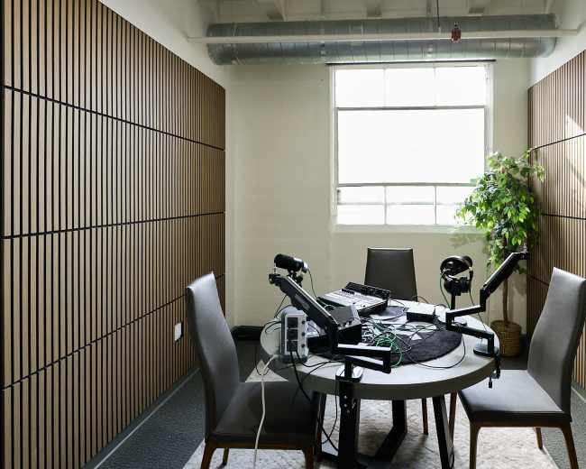 The image shows a small recording studio with a round table, two chairs, microphones, and audio equipment, featuring soundproofed walls and a window.