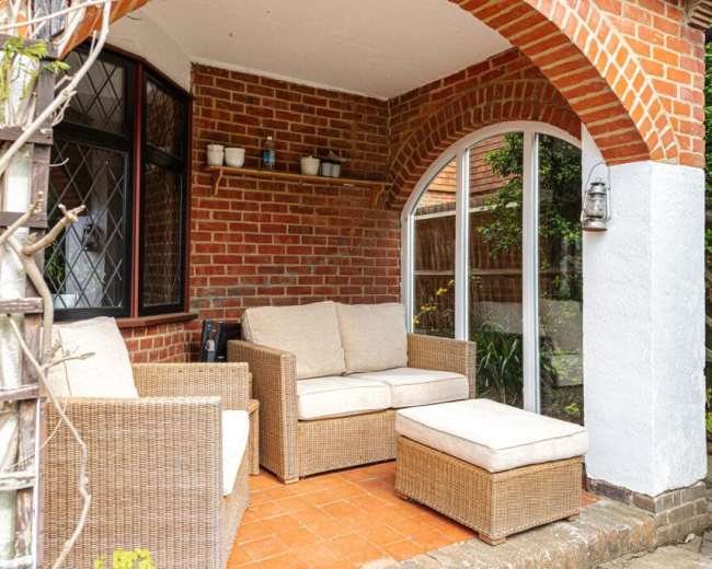 A cozy outdoor seating area with rattan furniture, including a sofa and an ottoman, set against a brick wall under an archway.
