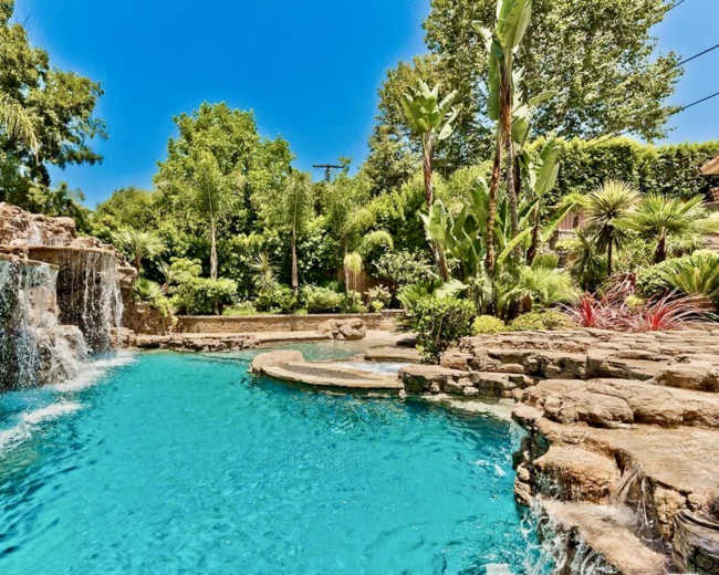 A clear blue swimming pool features a rock waterfall and lush tropical landscaping.