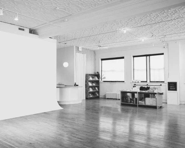 The image shows a spacious, well-lit studio with a white backdrop, large windows, and a shelving unit filled with various items.