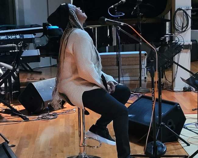 A musician sits on a stool in a recording studio, facing a microphone and a grand piano.