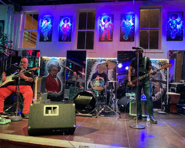 A band performs on stage in a venue adorned with colorful artwork while the audience observes.