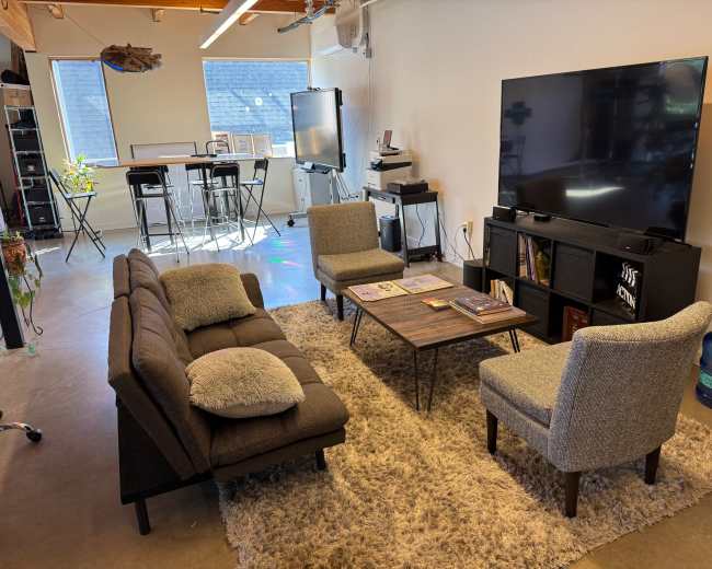 The image shows a modern lounge area with a brown sectional sofa, two grey armchairs, a coffee table with magazines, and a large television mounted on a black cabinet, all situated on a light-colored rug.