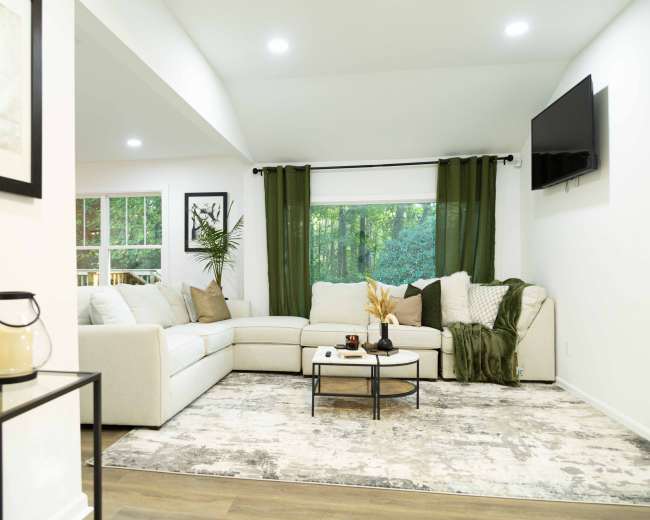The image shows a modern living room featuring a light-colored sectional sofa, a round coffee table, and large windows with green curtains, surrounded by decorative plants.