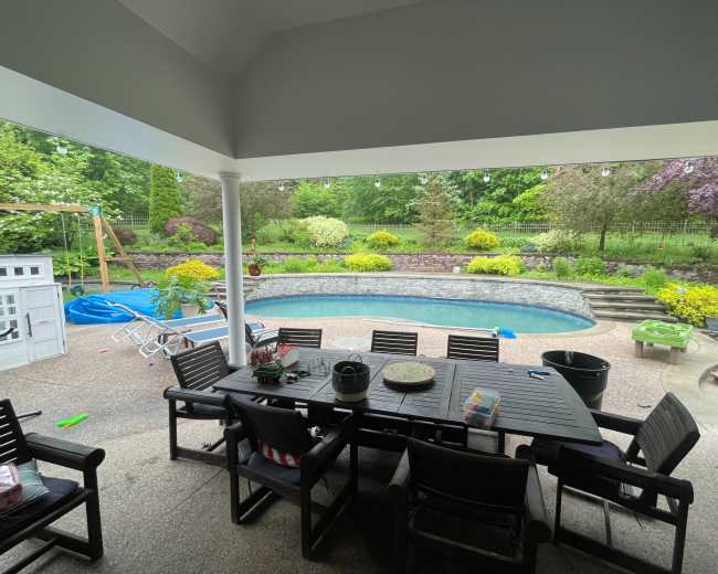 The image shows a covered outdoor patio area with a dining table, overlooking a pool surrounded by landscaped greenery and a playset in the background.