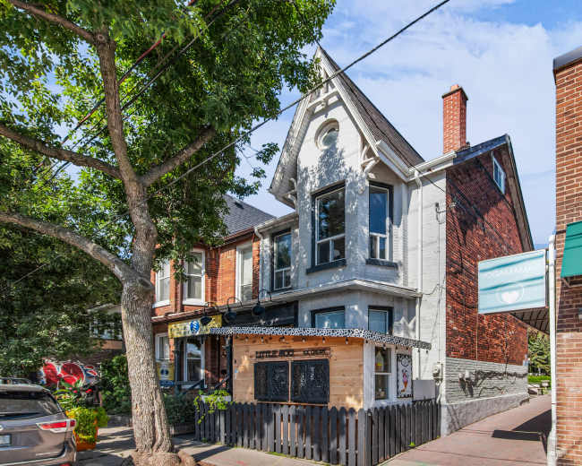 The image shows a two-story building with a peaked roof, featuring a mix of brick and gray siding, situated at the intersection of a street lined with trees and nearby commercial storefronts.