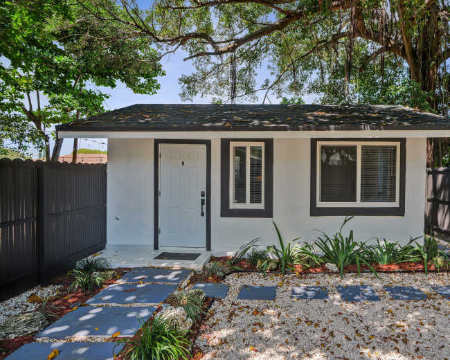 The image shows a small, modern house with a gray roof, white walls, and a stone pathway leading to the front door, surrounded by greenery and a black fence.