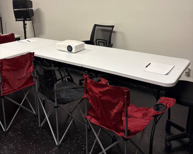 A rectangular table is set up in a stark room with several folding chairs and a projector placed in the center.