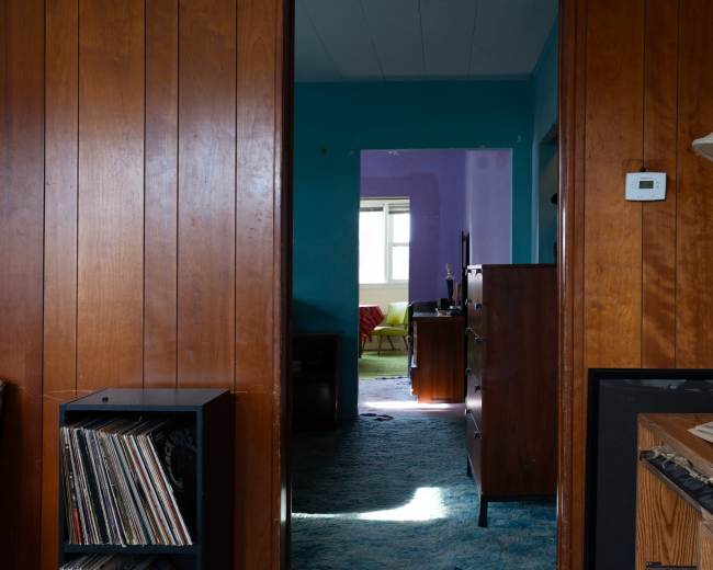 A doorway framed by wooden paneling opens into a brightly colored room with furniture visible in the background.