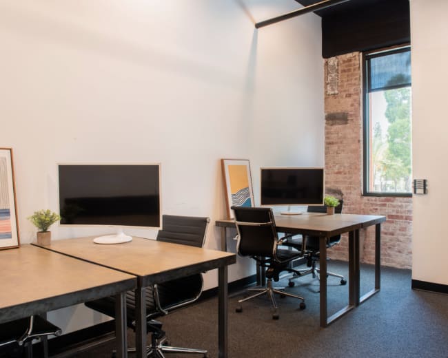 The image shows a modern office space with two desks, each equipped with a monitor, and a window providing natural light.
