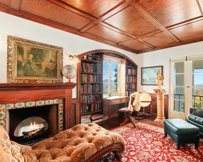The room features a wooden ceiling, a fireplace, a bookshelf, and large windows overlooking a scenic view.