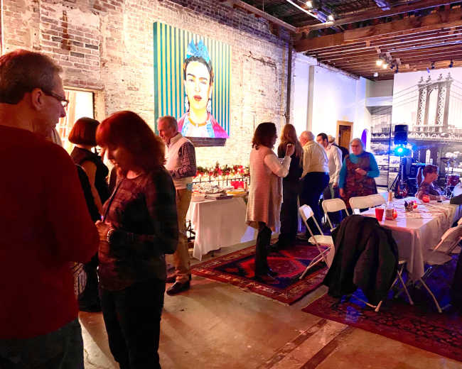 A crowded gallery space features people mingling around tables set with dishes, while a colorful portrait of a woman hangs on the brick wall.