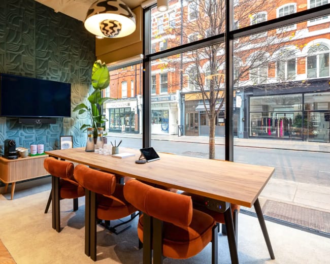 A modern conference room features a large wooden table surrounded by orange chairs, plants, and a wall-mounted television, with large windows overlooking a city street.