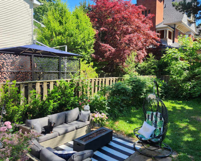 The image shows a landscaped backyard with a seating area featuring a sectional couch, a striped rug, and a hanging chair, surrounded by greenery and flowering plants.
