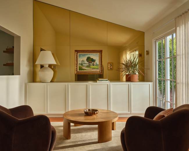 The image shows a modern living room featuring two brown chairs, a circular wooden coffee table, and a large mirror reflecting a painting and plants.