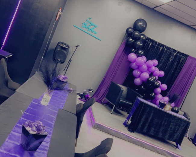 A decorated event space with a long table covered in a purple tablecloth, surrounded by black chairs, and a balloon arch in shades of purple and black in the background.