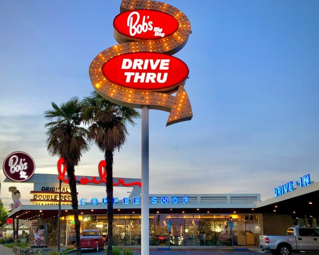 The image shows a retro-style diner sign for "Bob's Big Boy" with a drive-thru, set against the evening sky alongside the diner building featuring bright neon lights.