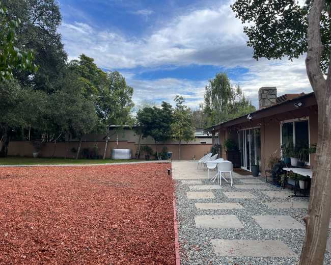 A patio area with stone pathways and white chairs is surrounded by a red mulch-covered yard and trees under a cloudy sky.