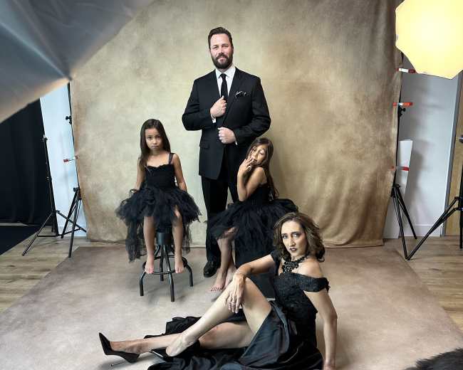 A man in a black suit stands with two girls in black dresses, while a woman in a black gown poses on the floor in front of them, all against a neutral backdrop.