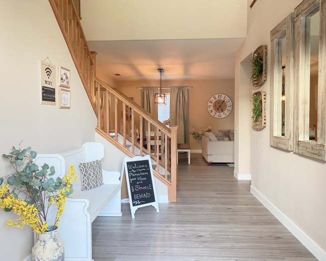The image shows a well-lit entryway with wooden stairs, a white bench, decorative wall art, and a chalkboard sign.