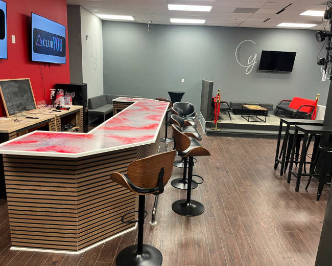 The image shows a modern lounge area with a bar, high tables, and seating arrangements, featuring a mix of red and neutral-colored walls.