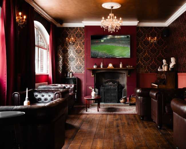 A stylish lounge features dark wood flooring, leather seating, and a large television displaying a sports event above an ornate fireplace.