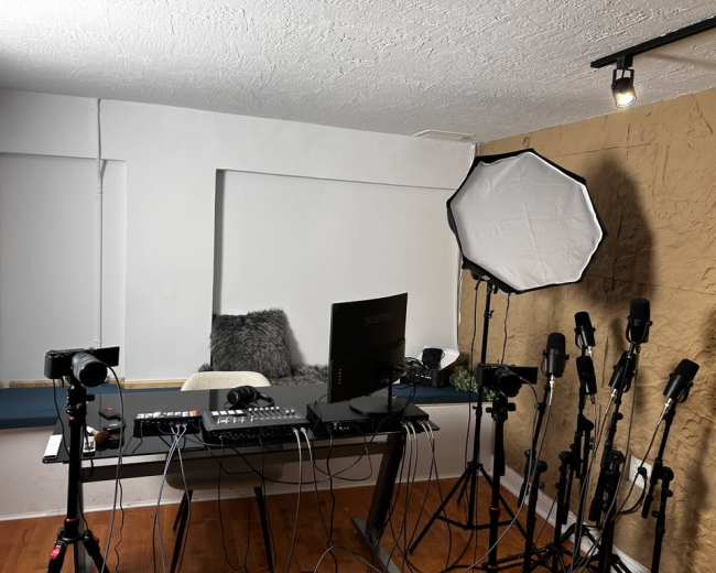 The image shows a recording studio setup featuring a large desk with a computer, multiple cameras, microphones on stands, and a softbox light.