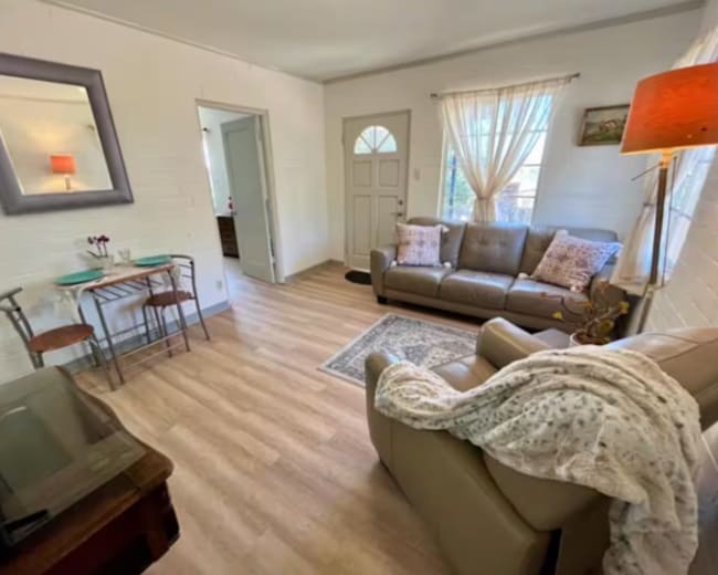 The image shows a light-filled living area with a brown leather sofa, a small dining table, and hardwood flooring.