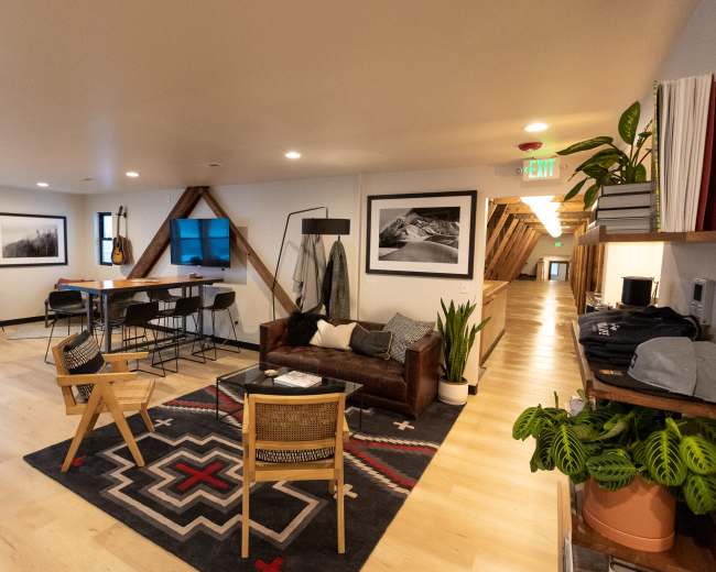 A modern open-concept lounge area featuring a leather sofa, wooden chairs, and a rug, with a dining space and hallway in the background.