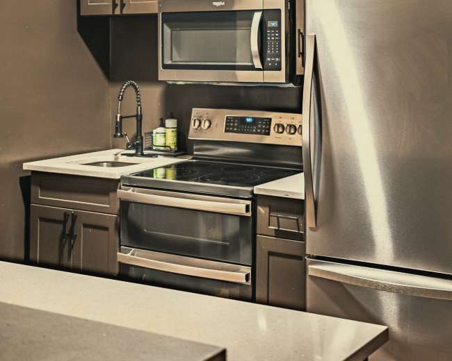 The image shows a compact kitchen featuring dark cabinets, a stainless steel refrigerator, a microwave above an electric stove, and a sink with a faucet.