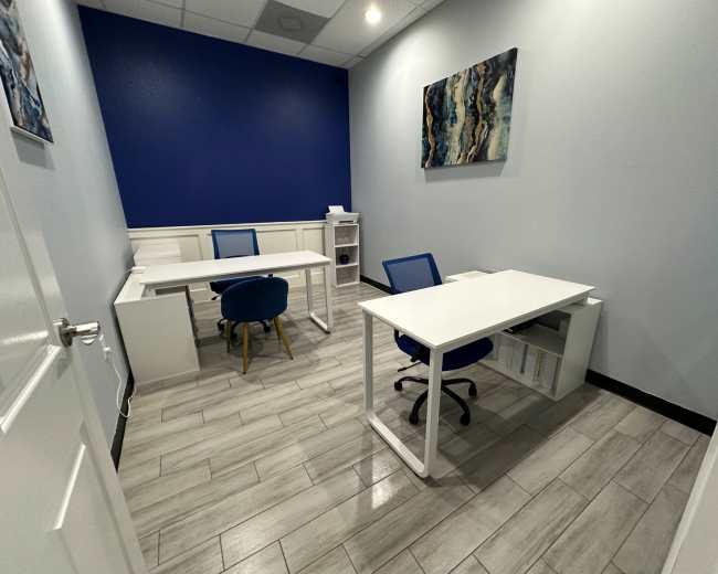 The image shows a small office with two desks, one blue chair, and a blue accent wall.