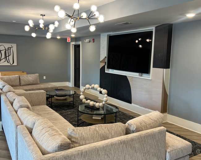 The image shows a modern lounge area featuring a sectional sofa, a circular coffee table, and a large wall-mounted television, with contemporary lighting fixtures overhead.