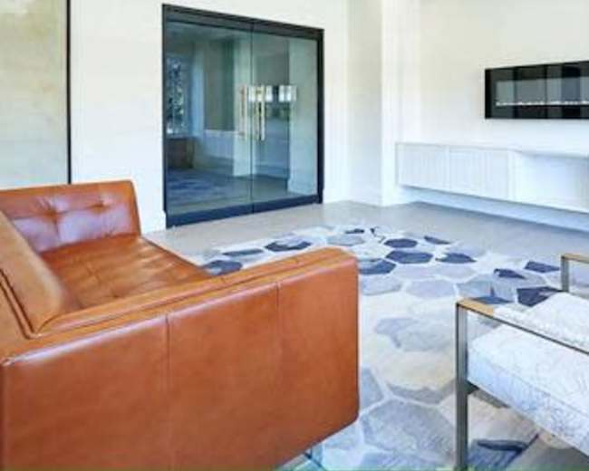 A modern, minimalist interior with a brown leather sofa, two white armchairs, and a patterned rug in a well-lit room featuring a glass door and a wall-mounted television.