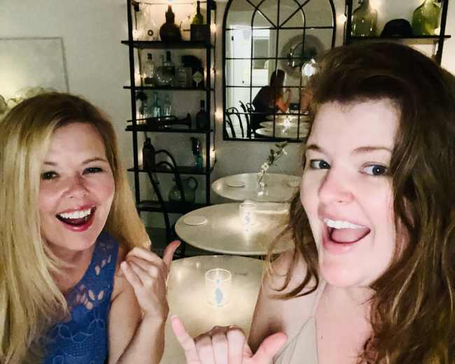 Two women pose for a selfie at a table in a restaurant, making hand gestures while smiling.