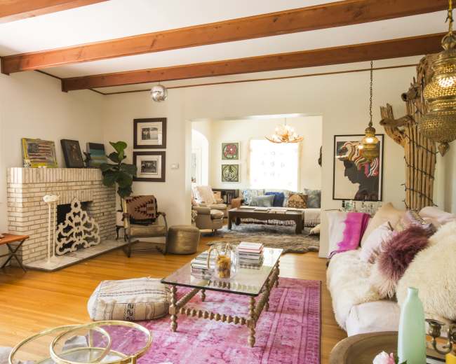 The living room features exposed wooden beams, a brick fireplace, a pink area rug, and various pieces of furniture including a sofa and a coffee table.