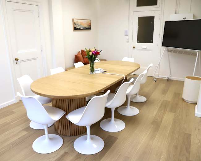 A modern meeting room features a large, oval wooden table surrounded by white, curved chairs, with a vase of flowers at the center.