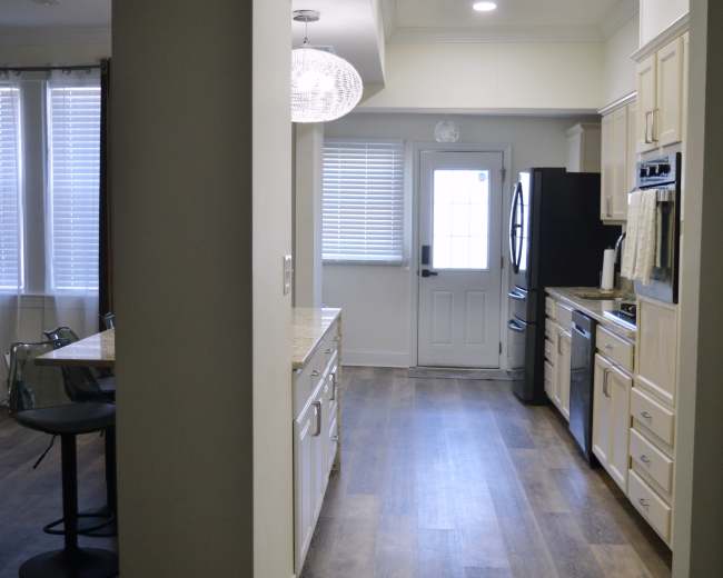 The image shows a modern kitchen with light-colored cabinets, a door leading outside, and a view of a dining area with a glass-top table in the background.