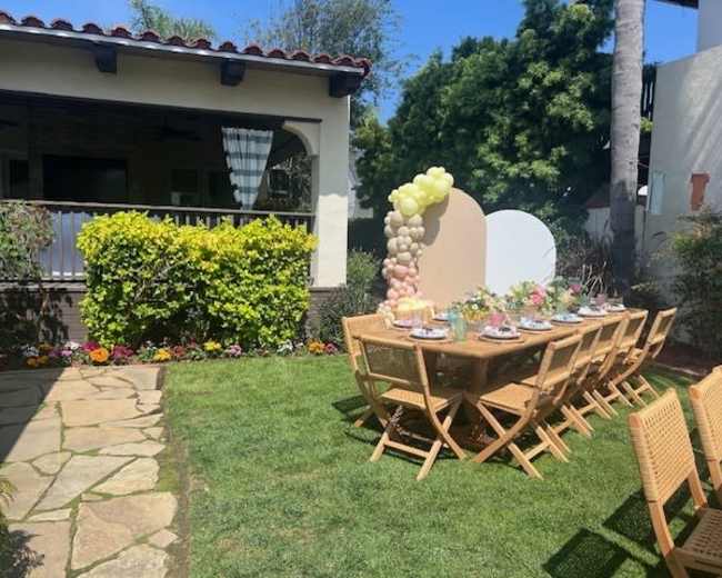 A long, wooden table set for a meal is placed on a grassy area surrounded by greenery and colorful flowers, with a house in the background.