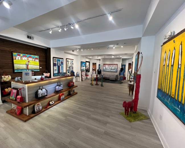 The image shows a bright, modern art gallery featuring various artworks and display shelves with colorful merchandise.