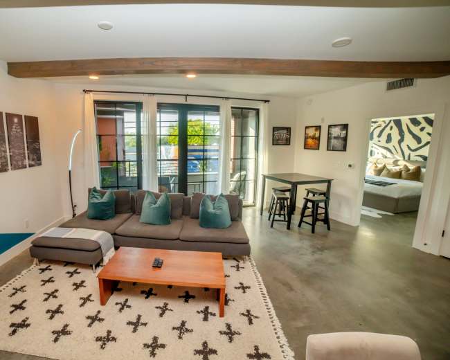 The image shows a modern living room with a sectional sofa, a coffee table, and a dining area in the background, featuring large windows and decorated walls.