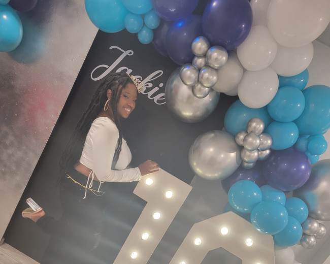 A young woman poses beside a large illuminated "16" surrounded by blue, silver, and white balloons.