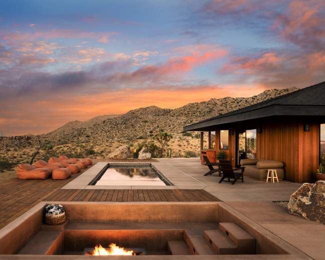 A modern home features a pool and fire pit, surrounded by desert landscapes and mountains under a colorful sunset sky.