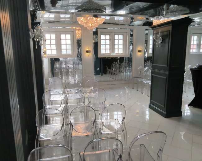 The image shows a modern event space with clear acrylic chairs arranged in rows, and large windows reflecting light from a chandelier above.