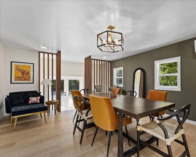 The image shows a modern dining area featuring a wooden table surrounded by orange and black chairs, with large windows and decorative wall elements.