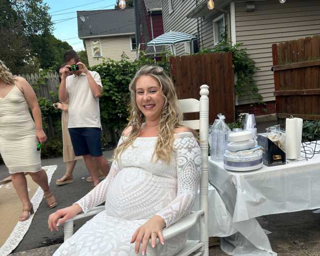 A pregnant woman in a white dress sits in a rocking chair at an outdoor gathering surrounded by decorations and party supplies.