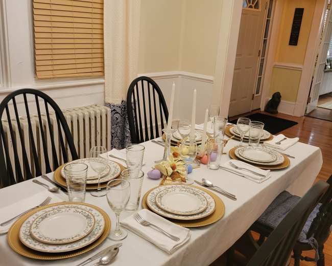 A dining table is set with plates, glassware, and cutlery, arranged neatly for a meal in a well-lit room with a doorway leading to another area.